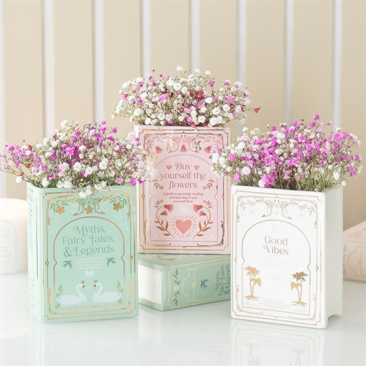 White ceramic book-shaped vase holding pink and white blooms, flanked by pastel book-style planters.
