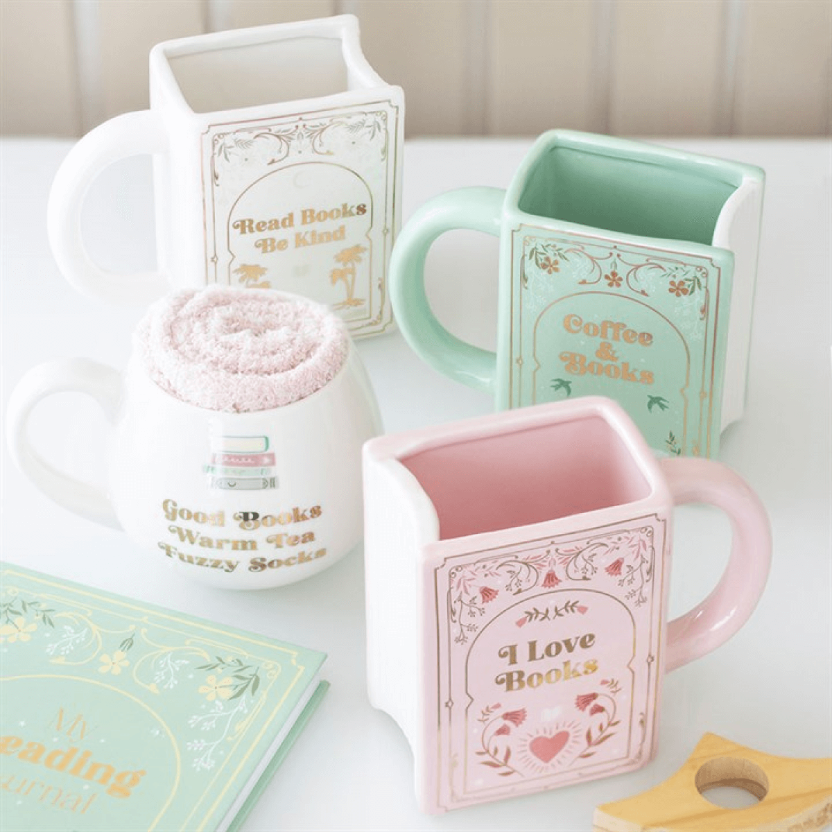 Read Books Be Kind white book-shaped ceramic mug with gold lettering, displayed among pastel book mugs.