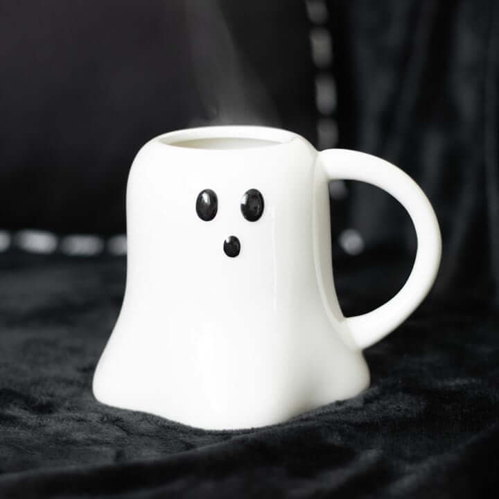 Ghost-shaped white ceramic mug for Halloween with black facial features and rising steam.