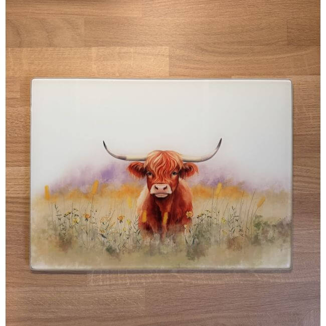Glass cutting board with a painted design of a Highland cow in a field on a wooden surface.