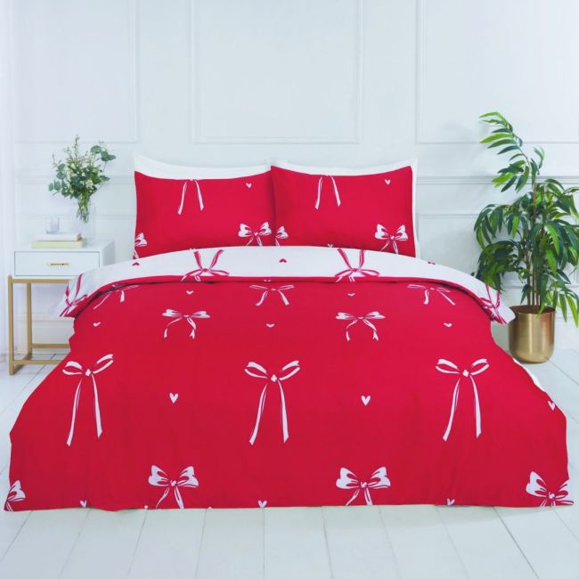 A red bedding set with a white bow and heart pattern, styled on a bed in a bright, modern bedroom with white paneled walls, a side table, and a potted plant.