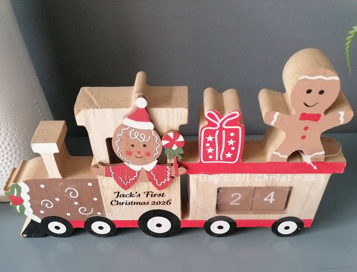 A wooden Christmas train decoration with gingerbread-themed details, including a Santa figure, a gingerbread man passenger, a red gift box, and sliding number blocks on the cargo car. The front car is personalized with the text 'Jack's First Christmas 2026'.