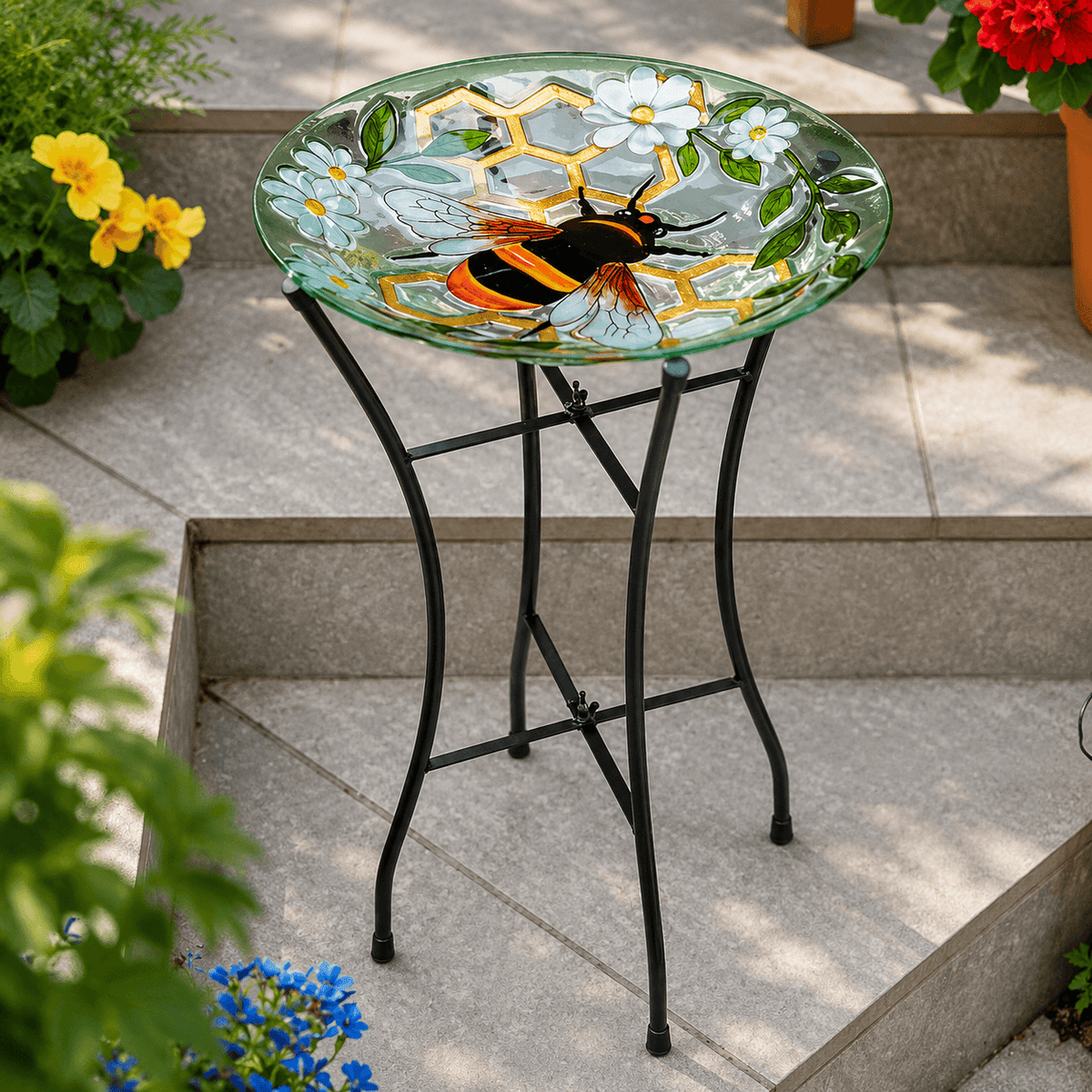 A round glass bird bath with a black metal stand, decorated with a bumblebee and white daisies on a green glass surface, set outdoors on stone steps with potted plants.