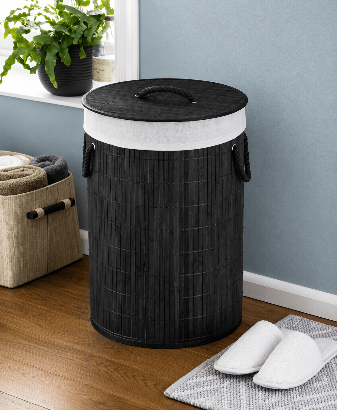 A black woven cylindrical laundry hamper with a white fabric liner and rope-style handles, shown in a styled room setting with a wood floor, blue wall, potted plant, folded towels, and slippers.