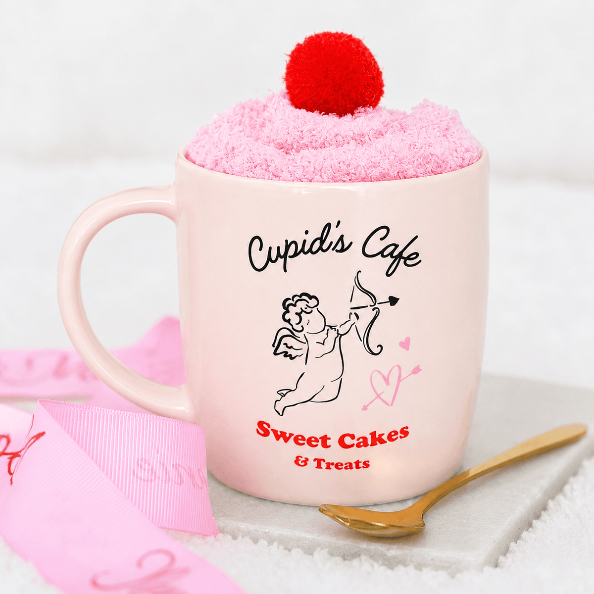 A light pink ceramic mug with 'Cupid's Cafe' and 'Sweet Cakes & Treats' printed on it, filled with fluffy pink frosting topped with a red pom-pom, styled on a white surface with a pink ribbon and gold spoon.