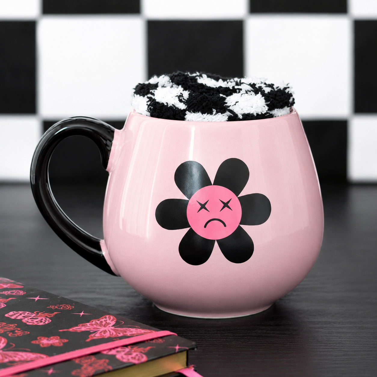A pink ceramic mug with a black handle and a black flower graphic featuring a pink sad face. The mug is filled with a black-and-white patterned beverage and sits on a dark tabletop with a black-and-white checkered background and a notebook in the foreground.