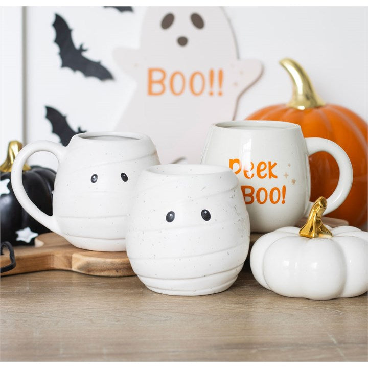 Three white mummy-shaped oil burners and wax warmers with black eyes, set among Halloween pumpkins and a Boo sign.