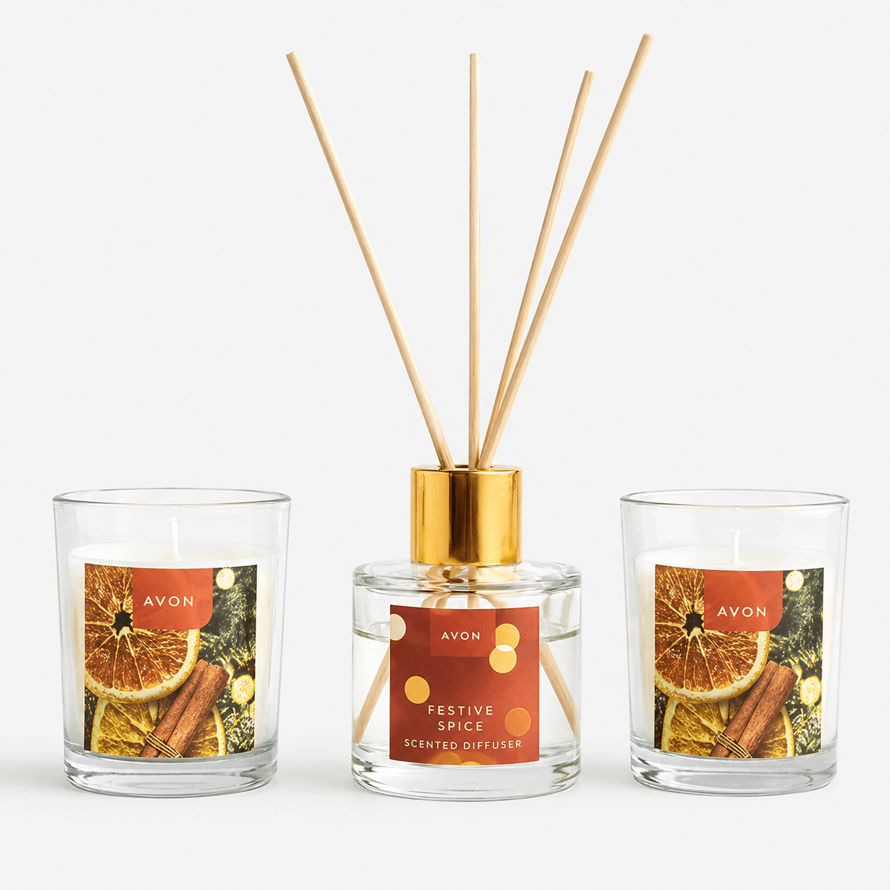 Three Avon scented home products on a light gray background: a candle in a clear glass cup with a citrus and cinnamon label, a clear glass reed diffuser bottle with a gold collar and wooden reeds, and another candle in a matching glass cup with a similar label.