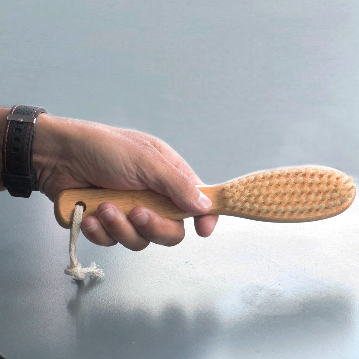 Beard brush with bamboo handle and boar hair bristles, held in a man's hand; rope loop at end.