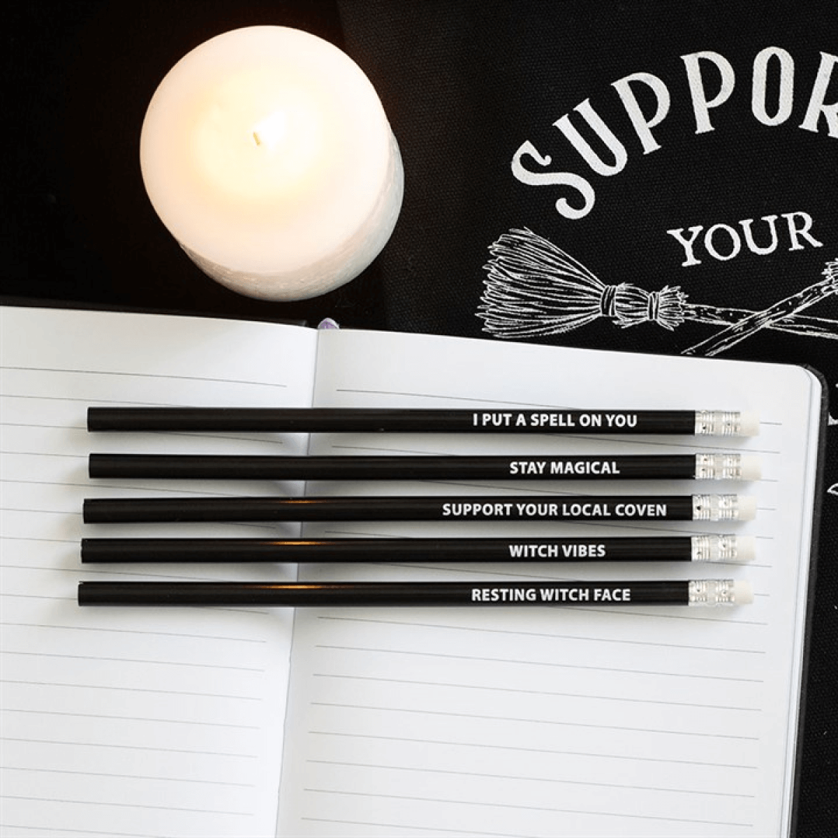 The Coven Club 5 Pack Witchy Pencils With Mystic Phrases displayed with a candle and a notebook.