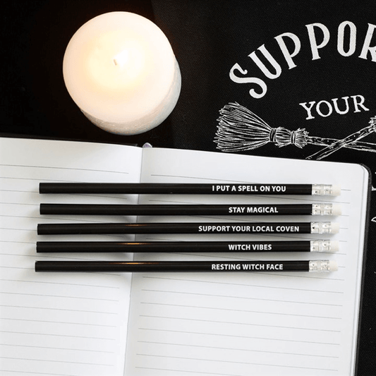 The Coven Club 5 Pack Witchy Pencils With Mystic Phrases displayed with a candle and a notebook.